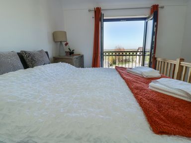 A bedroom with a bed covered in white and orange bedding with towels on top and a small dresser with a lamp and plant near an open door to a balcony at Bwlch Y Gwynt Bach in Llangefni