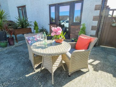 An outdoor patio with wicker chairs, a round wicker table, flowers on the table, and potted plants near a house at Bwlch Y Gwynt Bach in Llangefni