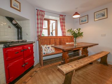 A dining area with a wooden table bench and bench seating a red stove and window with red check curtains at Ribby Farmhouse in Lerryn