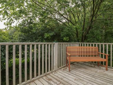 A wooden bench on a wooden deck with a railing overlooking green trees at Ribby Farmhouse in Lerryn