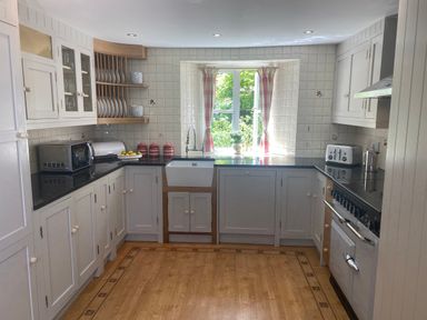 A kitchen with white cabinets black countertops a farmhouse sink and a window with pink check curtains at Ribby Farmhouse in Lerryn