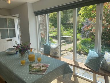 A dining area with a table covered in a blue polka dot tablecloth and windows with a garden view at Ribby Farmhouse in Lerryn