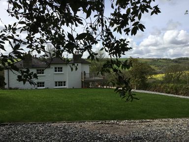 A white house with a porch overlooking a grassy yard and rolling hills in the background at Ribby Farmhouse in Lerryn