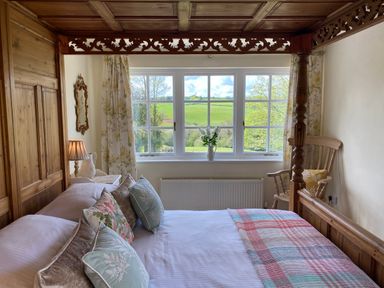 A bedroom with a wooden four poster bed and pillows next to a window with floral curtains and a wooden chair at Ribby Farmhouse in Lerryn