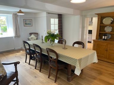 A dining room with a large table covered by a polka dot tablecloth six wooden chairs a wooden cabinet with plates and wine bottles and a vase with flowers on the table at Ribby Farmhouse in Lerryn