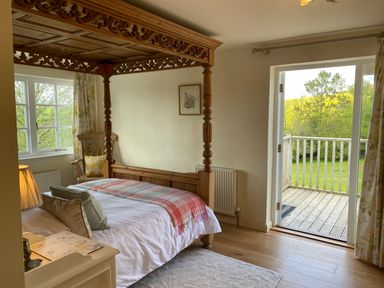 A bedroom with a wooden four-poster bed a wooden chair by a window a bedside table with a lamp and an open door leading to a balcony at Ribby Farmhouse in Lerryn