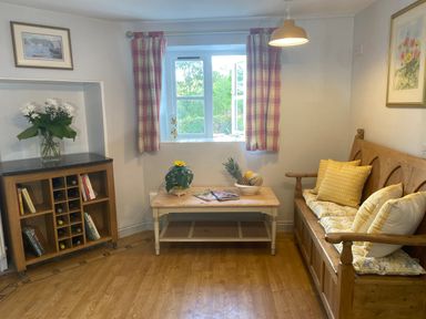 A sitting room with a wooden bench and cushions a wooden coffee table with plants and magazines a wooden wine rack with books and flowers on top at Ribby Farmhouse in Lerryn