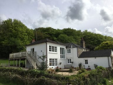 A white two-story house with multiple windows and a wooden deck surrounded by trees and greenery at Ribby Farmhouse in Lerryn