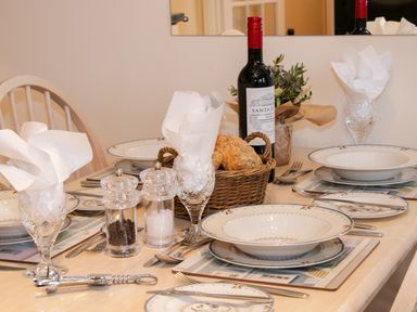 A dining table set with plates, cutlery, crystal glasses with white napkins, a basket of bread, salt and pepper shakers, a bottle of wine, and a small flower arrangement at Cream Door Cottage in Kidderminster