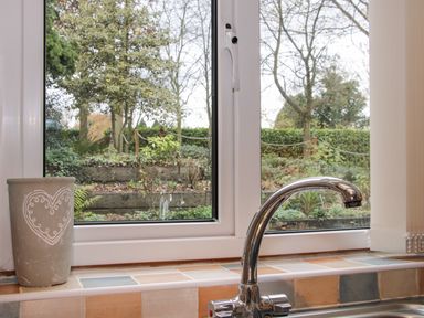 A kitchen window with a metal faucet and a pot on the windowsill overlooking a garden at Cream Door Cottage Kidderminster