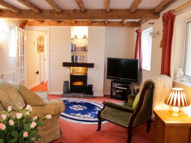 A living room with wooden beams on the ceiling a TV a fireplace an armchair a sofa a round blue rug and flowers on a table at Cream Door Cottage in Kidderminster