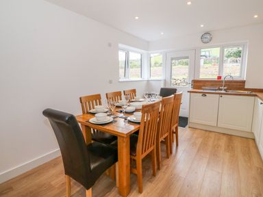 A dining area with a wooden table set for six with chairs next to a kitchen with white cabinets and a door with windows at Llwyd Coed Canol in Llanllyfni near Penygroes