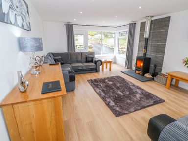A living room with a gray sofa a wood burning stove a wooden cabinet and large windows at Llwyd Coed Canol in Llanllyfni near Penygroes