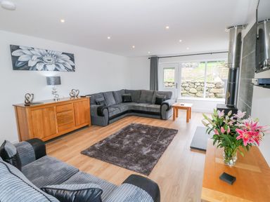 A living room with gray sofas a wooden cabinet with decorations a glass vase with flowers and a wall-mounted stove at Llwyd Coed Canol in Llanllyfni near Penygroes