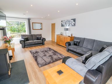 A living room with gray sofas a wooden cabinet a small wooden table a fireplace and a large window at Llwyd Coed Canol in Llanllyfni near Penygroes