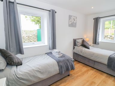 A bedroom with two single beds, gray bedding and pillows, two windows with gray curtains, a bedside table with a lamp, and a wall flower painting at Llwyd Coed Canol in Llanllyfni near Penygroes