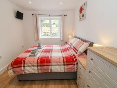 A bedroom with a double bed with red and beige plaid bedding a window and wooden dressers at Llwyd Coed Canol in Llanllyfni near Penygroes
