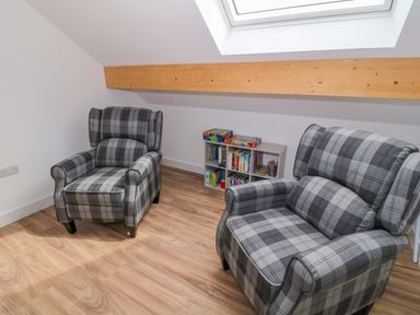 A room with two plaid armchairs a small bookshelf with books and board games under a skylight window at Llwyd Coed Canol in Llanllyfni near Penygroes