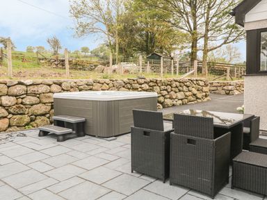 An outdoor patio with a hot tub and a glass table with six wicker chairs surrounded by a stone wall at Llwyd Coed Canol in Llanllyfni near Penygroes