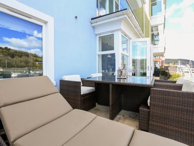 An outdoor patio with brown cushioned loungers wicker dining chairs and a glass table at Quayside Dart Marina in Dartmouth