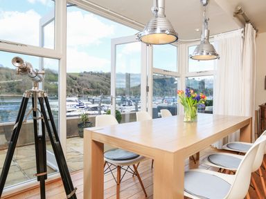 A dining area with a wooden table and white chairs next to large windows overlooking a marina with boats at Quayside Dart Marina in Dartmouth