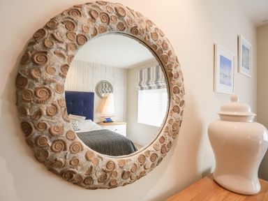 A round mirror with a shell patterned frame reflecting a bedroom with a bed a bedside table a lamp and a window at Quayside Dart Marina Dartmouth