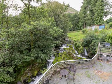 A patio with metal chairs and table overlooking a river with rocks and trees at Dyfi Cottage in Dolgellau