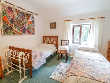 A bedroom with two single beds a wooden chair with folded towels a window with white curtains and a colorful wall hanging at Dyfi Cottage in Dolgellau