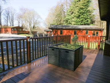 An outdoor wooden deck with a rattan table and railing overlooking wooden cabins and trees at 40 Skiptory Howe in Troutbeck Bridge