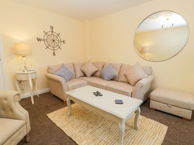 A living room with a beige sofa set and cushions a white coffee table with remotes and a coaster a side table with a lamp and decor a round wall mirror and a compass wall art at Valiant House in Scarborough