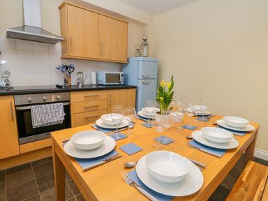 A kitchen with a wooden dining table set for six with white plates bowls wine glasses and a plant and light wood cabinets a blue refrigerator and microwave at Valiant House Scarborough