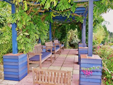 An outdoor seating area with wooden benches under a blue pergola surrounded by greenery at Tudor House in Symonds Yat
