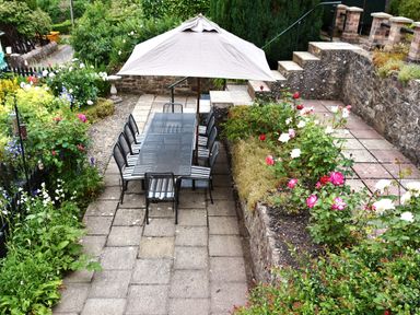 A garden patio with a metal table and chairs under a large umbrella surrounded by flower beds at Tudor House in Symonds Yat