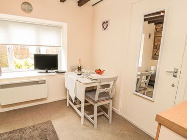 A small dining area with a table, two chairs, a bottle of wine, a bowl of fruit, a television on the window sill, a mirror on the door at Robin Cottage in Morpeth