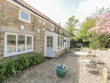 A stone exterior of a house with white framed windows a white door a small patio area with a white metal table and chairs and garden plants at Robin Cottage in Morpeth