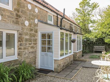 Stone building with a light blue door labeled Robin and large windows with hanging flower baskets and a garden bench at Robin Cottage in Morpeth
