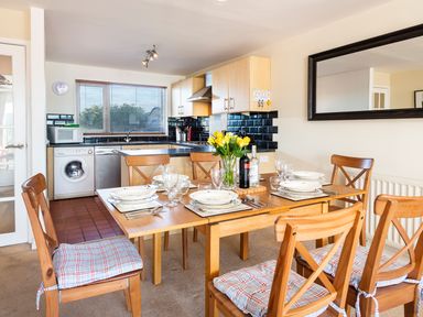 A dining area with a wooden table set for six and an open kitchen with appliances at 164 Cumber Close in Malborough