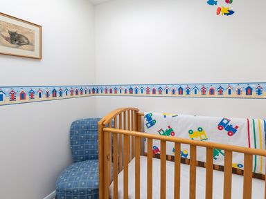 A nursery room with a wooden crib, blue upholstered chair, framed picture on the wall, and a wallpaper border with beach huts at 164 Cumber Close in Malborough