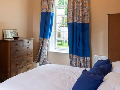 A bedroom with a wooden chest of drawers a bed with blue pillows and patterned curtains near a window at The Old School in Gortin