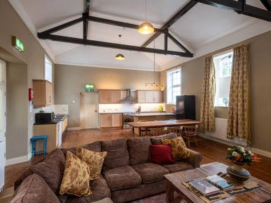 A living area with brown sofas and pillows a wooden coffee table with books and a vase an open kitchen with wooden cabinets a dining table with chairs and windows with curtains at The Old School in Gortin