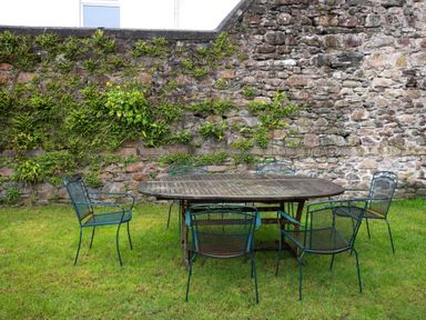 An outdoor area with a wooden table surrounded by six metal chairs on grass near a stone wall with plants at The Old School in Gortin