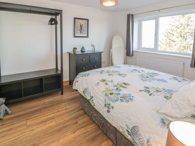 A bedroom with a bed with floral bedding a black chest of drawers a standing mirror a window and a black open wardrobe at Isallt in Rhostryfan near Bontnewydd Gwynedd