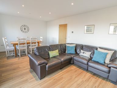 A living room with a brown leather sectional sofa with cushions a wooden dining table with six white chairs and a wall clock at Isallt in Rhostryfan near Bontnewydd Gwynedd