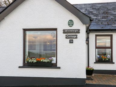 The exterior of Woodleigh Cottage with windows displaying flower boxes and a sign with the cottage name and number 76 at Ballycastle
