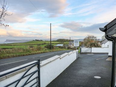 A driveway and paved area next to a white wall and gate with a road and green fields in the background at Woodleigh Cottage in Ballycastle