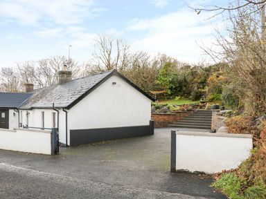 A driveway with a white house on the left and steps leading to a garden with greenery and a picnic table at Woodleigh Cottage in Ballycastle