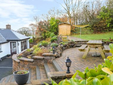 A garden with a patio area with a wooden picnic table a small shed and a white house with a black roof at Woodleigh Cottage in Ballycastle