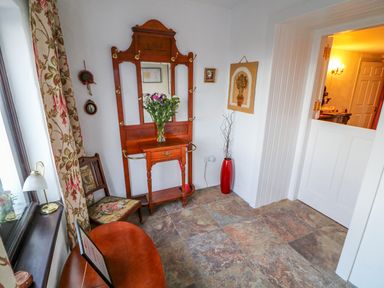 A small room with a wooden hall stand with a mirror a chair a red vase with branches and a wooden table by a window at Woodleigh Cottage in Ballycastle