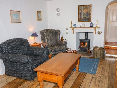 A sitting room with two armchairs a wooden coffee table a wood stove with fire and wooden floors at Woodleigh Cottage in Ballycastle