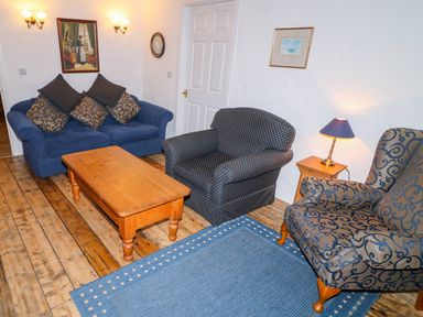 A living room with sofas a wooden coffee table a patterned armchair a wooden side table with a lamp and framed pictures on white walls at Woodleigh Cottage in Ballycastle